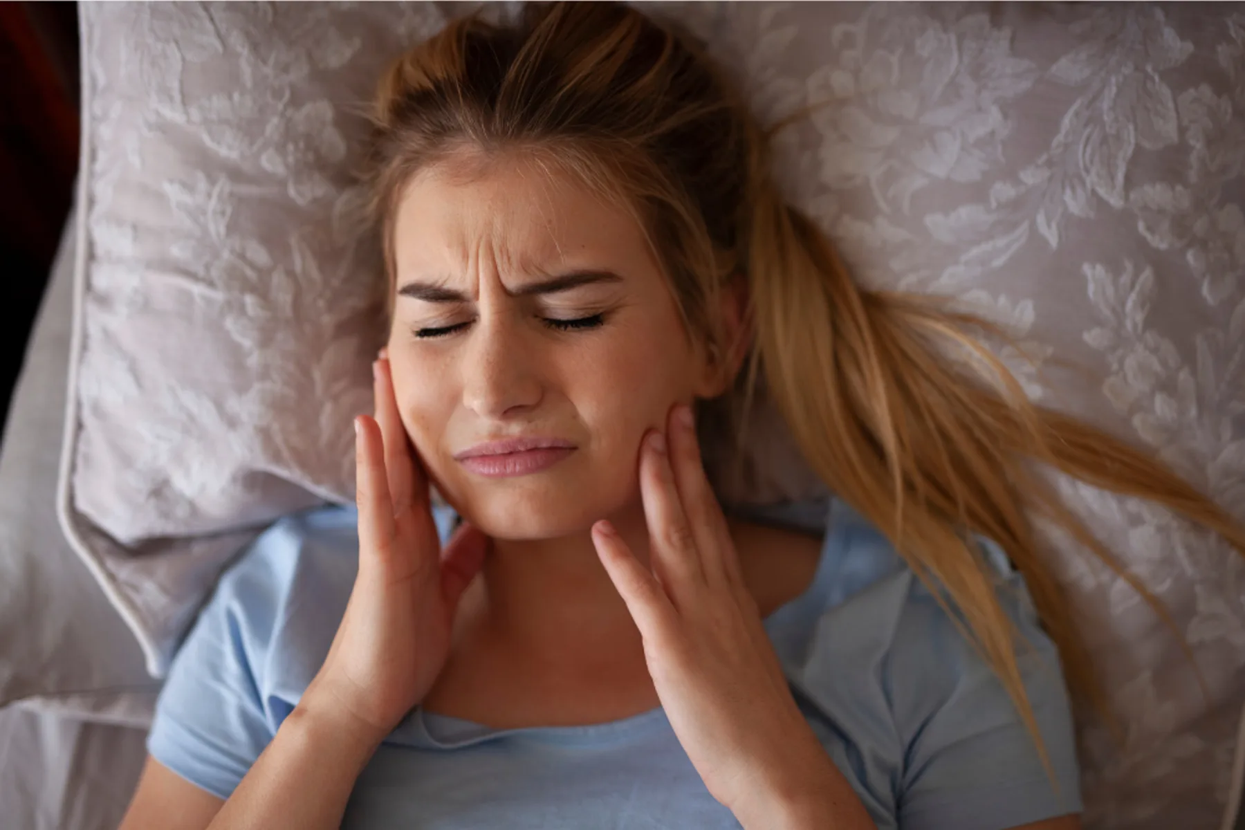 Woman lying in bed holding her jaw with morning jaw pain caused by bruxism